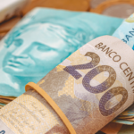 Notas de 200 e 100 reais em close-up, com moedas ao fundo.